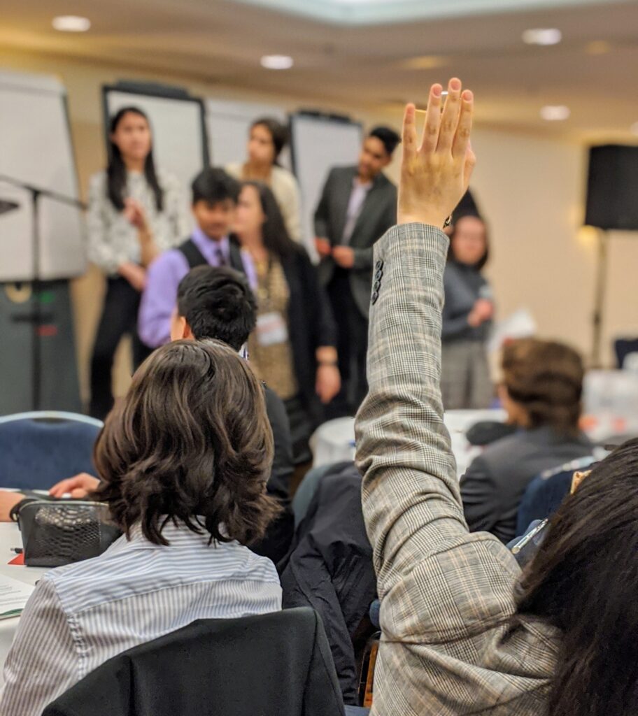 A photo of a group in session. The view is from the audience behind a person with their hand raised to speak.